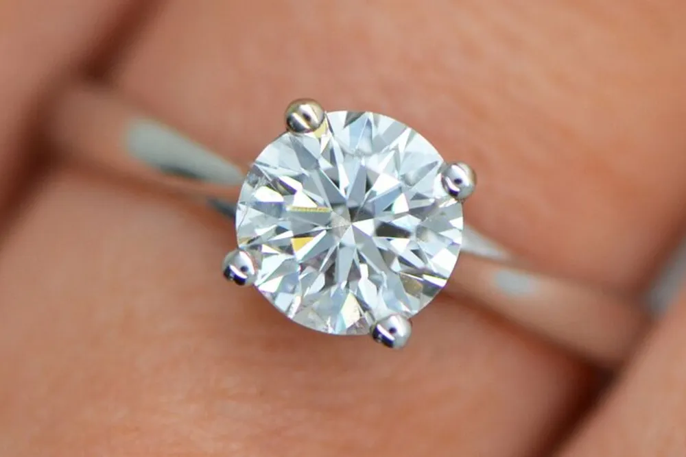 The image displays a close-up of a round-cut diamond engagement ring on a persons finger