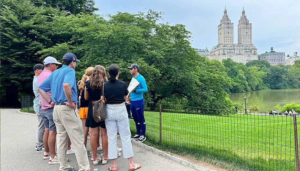 A group of people is gathered in a park possibly undergoing a guided tour with a scenic backdrop featuring a lake and towering buildings