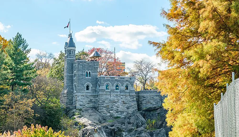 The image shows a small stone castle with a tower and ramparts set against a backdrop of trees displaying autumn foliage under a clear blue sky