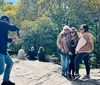 A person is taking a photo of three individuals posing together on a sunny day in what appears to be a park with trees and other people in the background