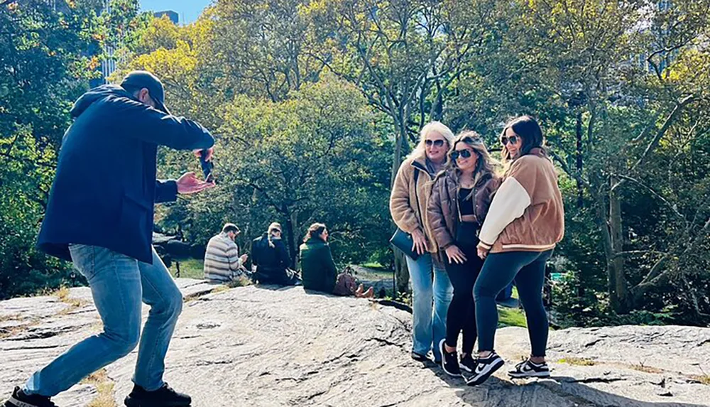 A person is taking a photo of three individuals posing together on a sunny day in what appears to be a park with trees and other people in the background