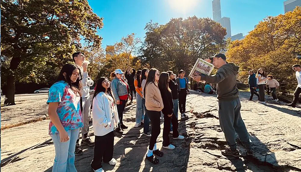 A group of people are attentively listening to a person who is holding a newspaper and gesturing during what appears to be a guided tour in an outdoor setting with rocky ground and trees against a backdrop of high-rise buildings