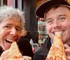 Two people are smiling at the camera while biting into large slices of pizza