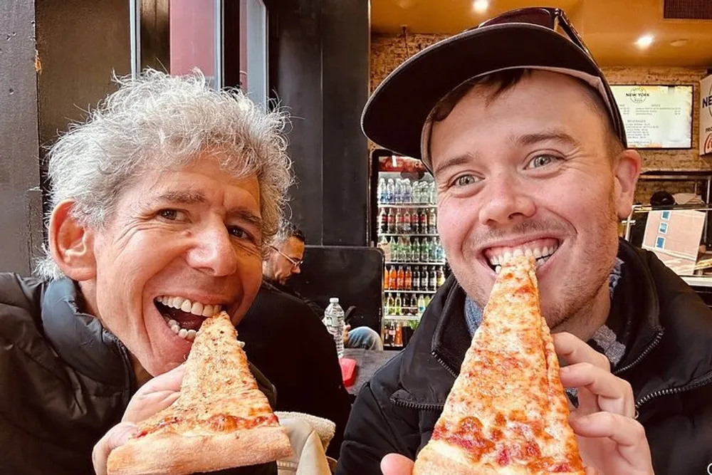 Two people are smiling and holding up large slices of pizza to their mouths as if taking a bite in a casual dining setting