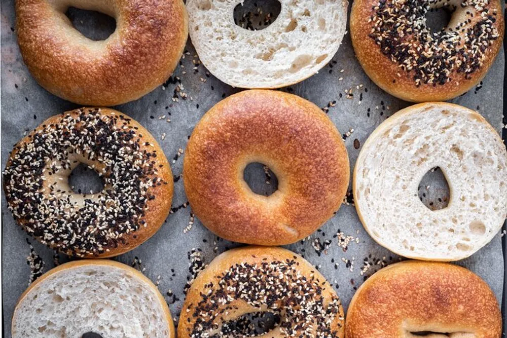 The image shows an assortment of several fresh bagels some whole and some sliced with different toppings like sesame seeds displayed on a parchment-lined flat surface