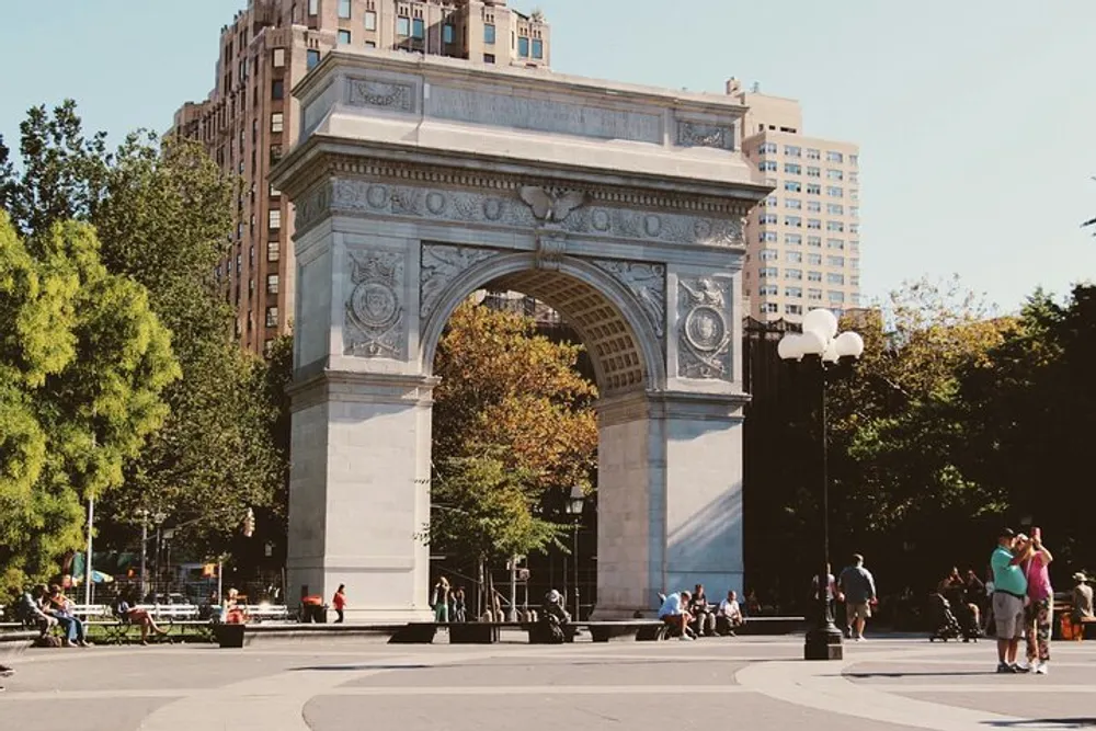 A large ornate arch stands prominently in a bustling urban park with people walking and enjoying the sunny weather