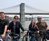 Four individuals pose with their bicycles on a waterfront with the Brooklyn Bridge in the background