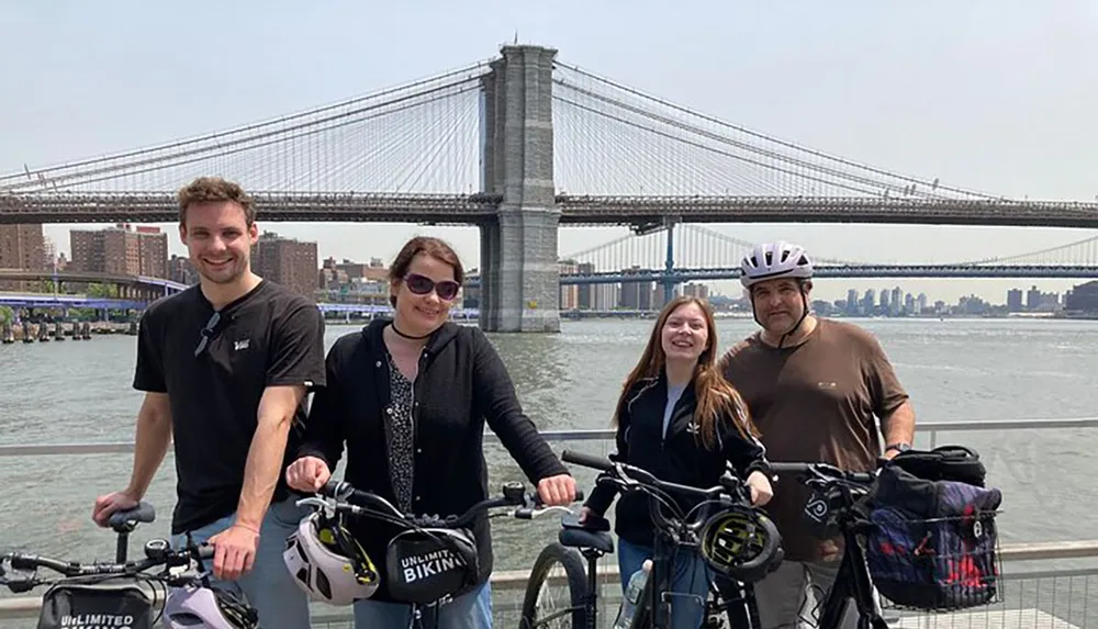 Four individuals pose with their bicycles on a waterfront with the Brooklyn Bridge in the background