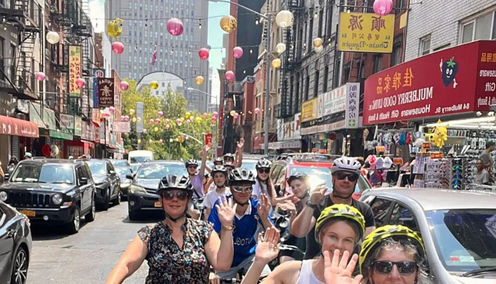 A group of cyclists wearing safety helmets is posing and waving to the camera on a street adorned with colorful lanterns indicating a vibrant urban environment possibly in a Chinatown district