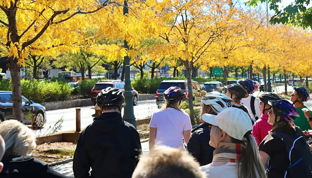 A group of people wearing bike helmets are gathered together on a sunny day with vibrant autumn foliage in the background