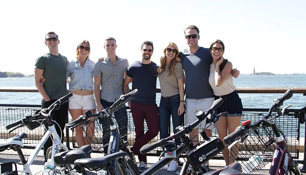 A group of seven smiling people is posing for a photo behind a row of parked bicycles with a body of water and a distant island in the background