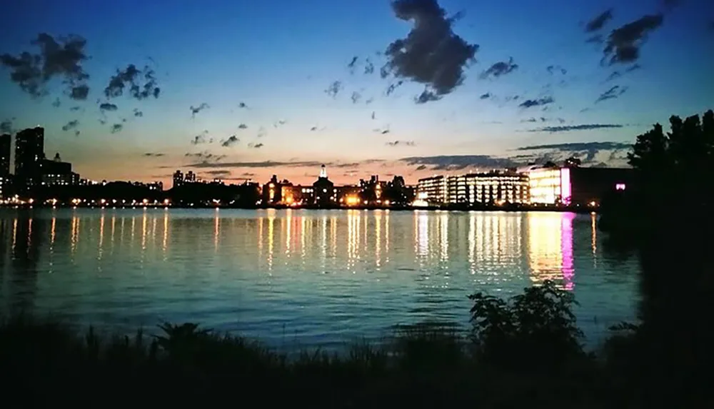 The image captures a tranquil urban twilight scene where the silhouettes of buildings and a spectrum of lights are reflected upon the calm surface of a body of water against a subtly lit evening sky