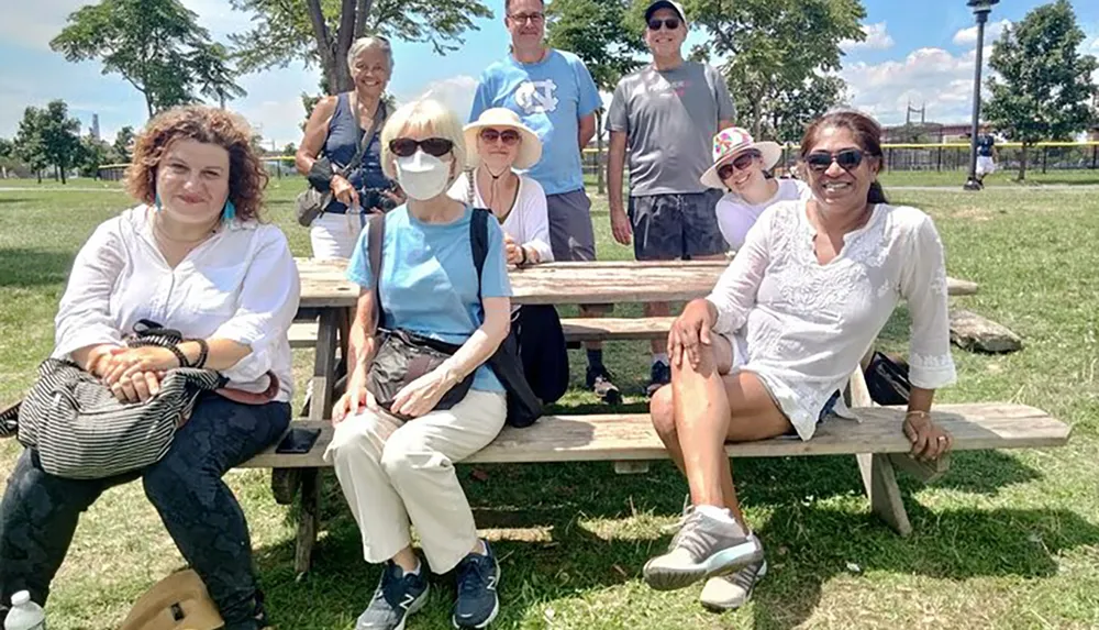 A group of eight people with diverse ages is casually posing for a photo while sitting and standing around a wooden picnic table in a sunny park setting