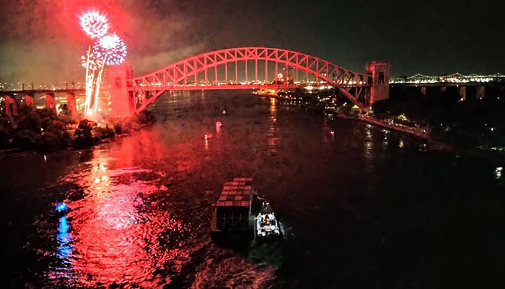 A nighttime display of fireworks over a lit-up arch bridge with a boat on the water below adds a festive atmosphere to the urban scene