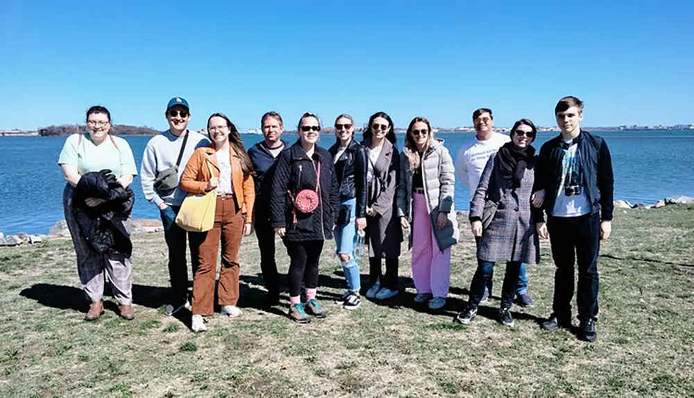 A group of people poses for a photo standing on grass near a body of water under a clear blue sky