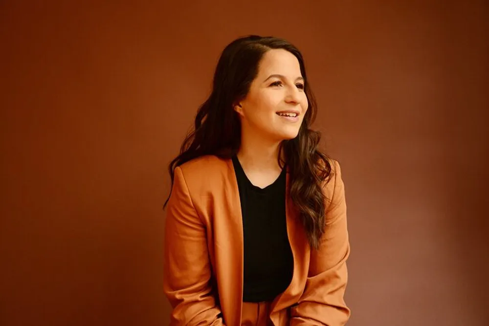 A smiling woman wearing a tan blazer looks off to the side against a brown background