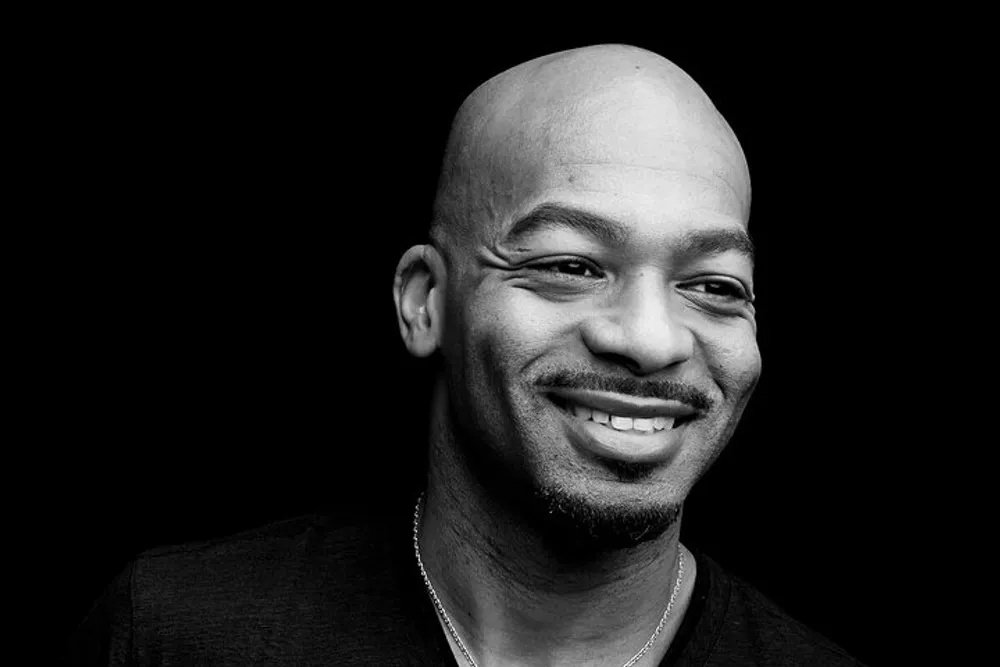 The image shows a smiling bald man with a goatee photographed in black and white against a dark background