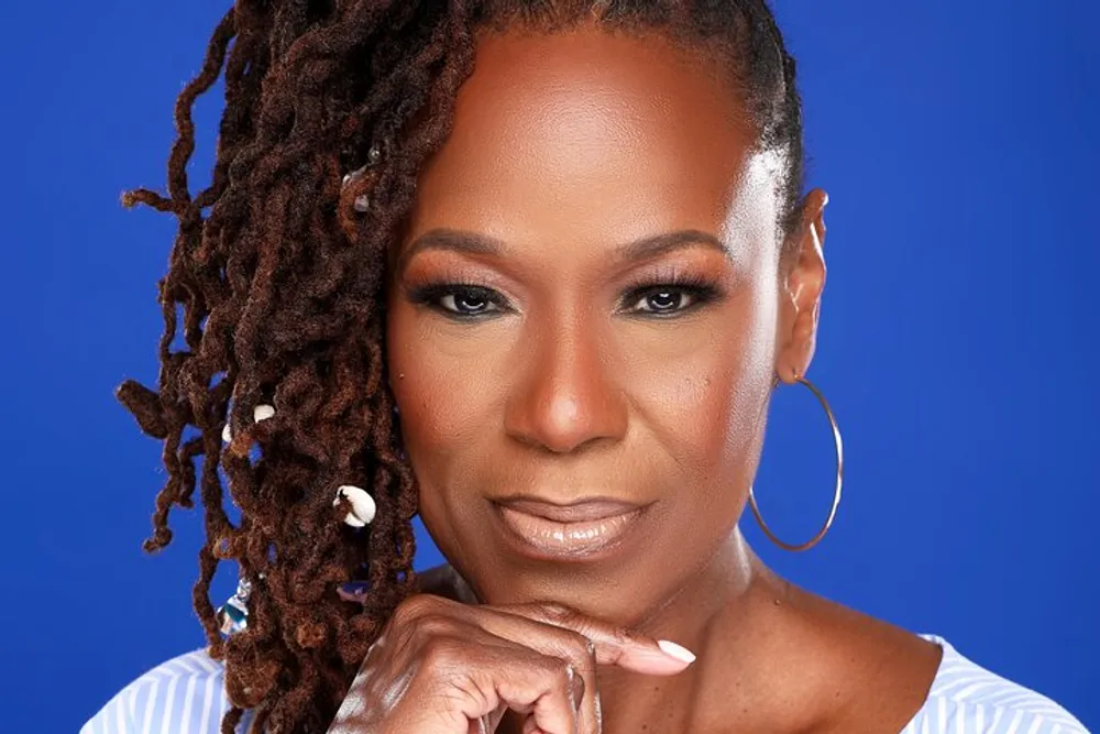 A portrait of a confident woman with shoulder-length dreadlocks wearing hoop earrings and a pinstripe garment set against a blue background