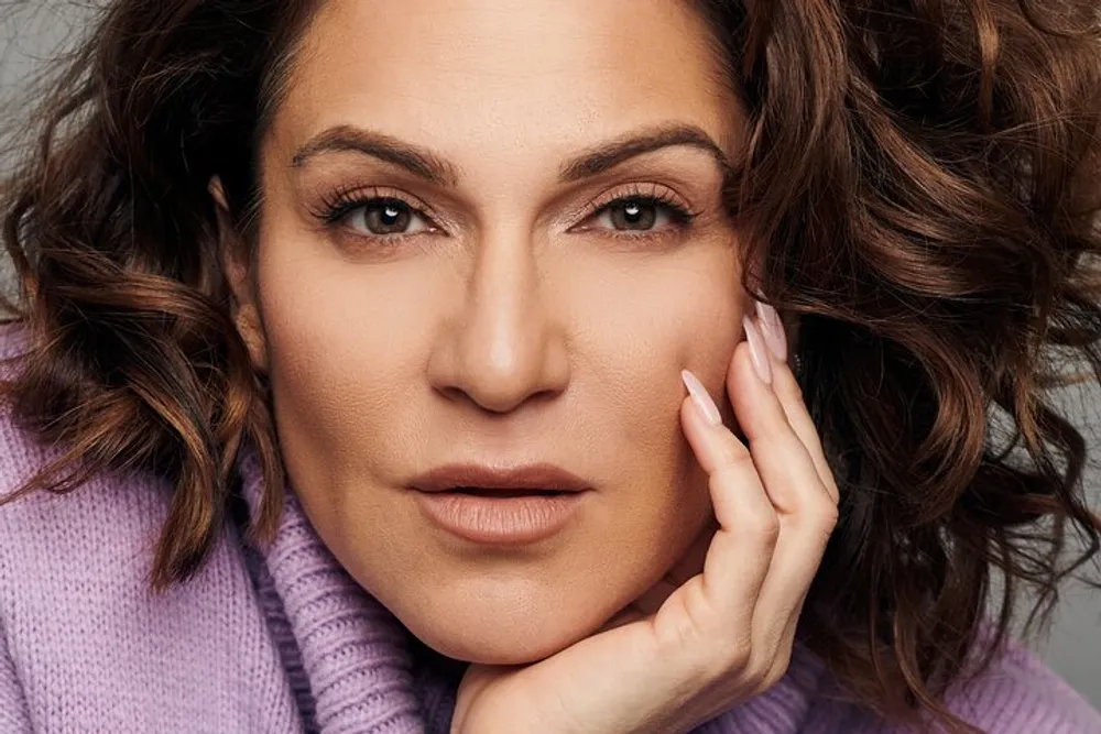 The image shows a close-up of a woman with wavy brown hair and striking features wearing a lilac top and resting her hand gently on her cheek exuding confidence and poise