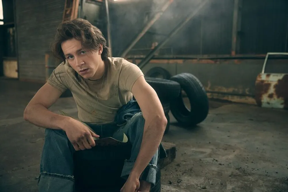 A person is crouching in a dimly lit garage-like setting with scattered car parts looking thoughtfully at the camera