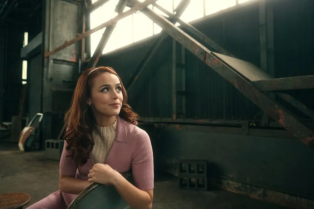 A smiling woman in a pink outfit is seated and looking to her left in an industrial setting with sunlight filtering through a window