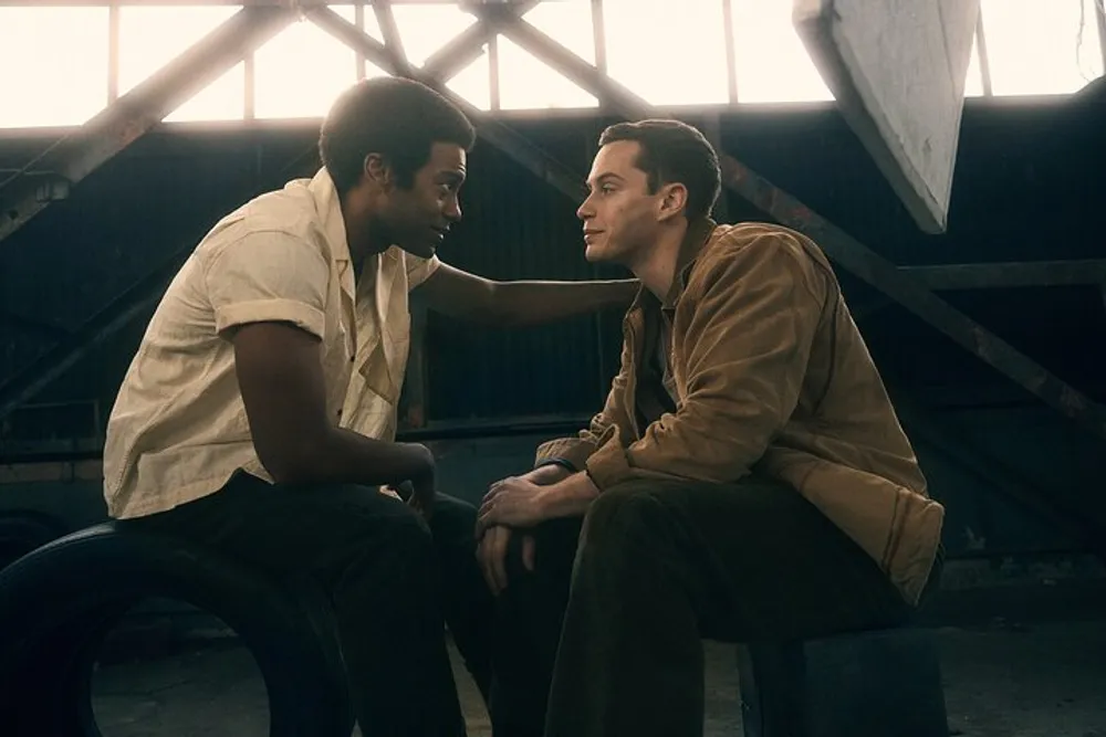 Two men are sharing an intimate moment in a dimly lit industrial setting with one gently placing his hand on the others shoulder while they gaze into each others eyes