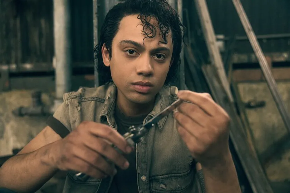 A young person with curly hair looks intently at a metallic object in their hands while sitting in a workshop-like setting