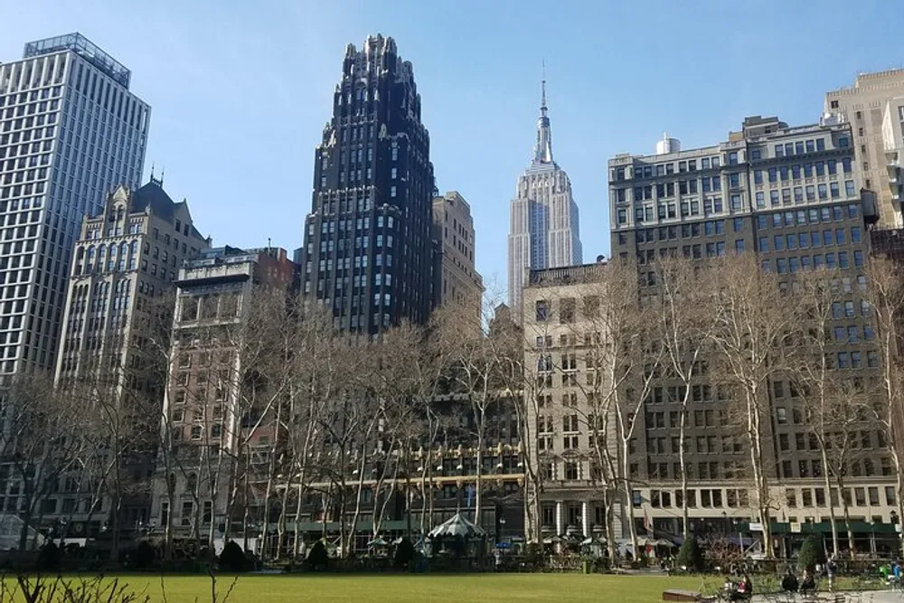 The image shows a view of a park in an urban setting with the Empire State Building towering in the background amidst other high-rise buildings under a clear blue sky