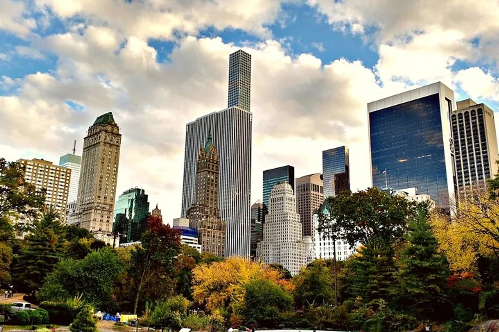 The image features a striking contrast between the vibrant autumn foliage of a park and the towering skyscrapers of a dense urban skyline under a partly cloudy sky