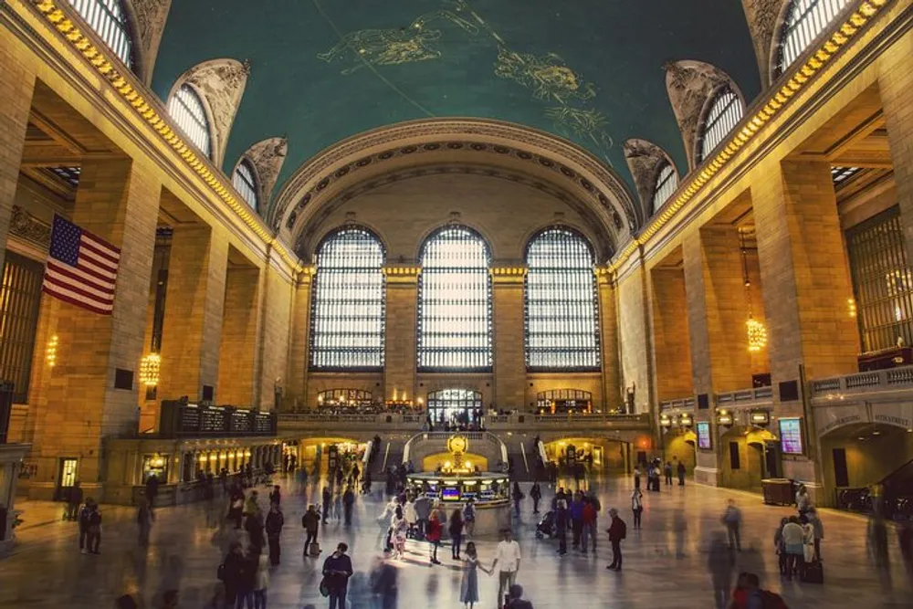 The image shows the bustling interior of Grand Central Terminal with its iconic celestial ceiling large arched windows American flag and people in motion throughout the concourse