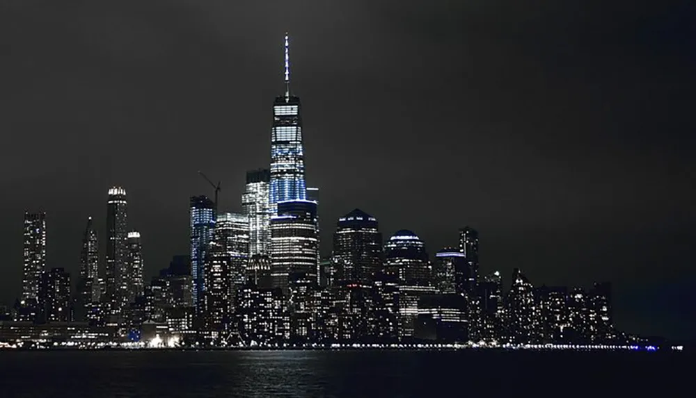 The image shows a nighttime skyline of a modern city with illuminated skyscrapers dominating the scene reflecting off the water in the foreground