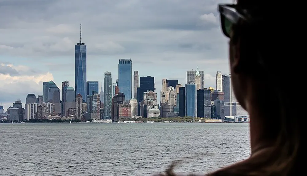 A persons silhouette in the foreground looks towards a cloudy Manhattan skyline featuring the One World Trade Center