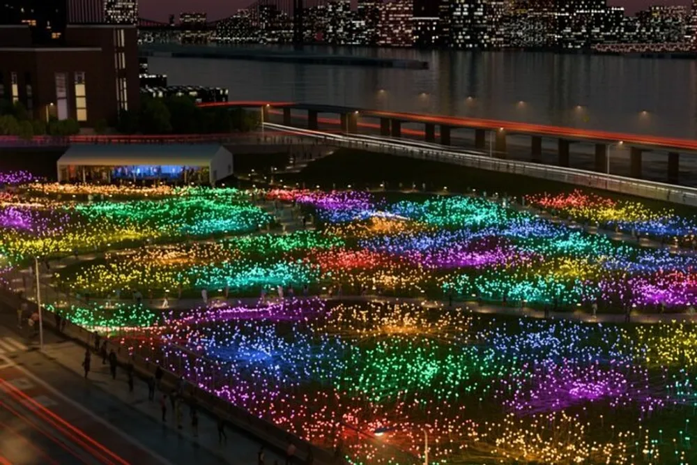 A vibrant display of multicolored lights blankets an outdoor field at night with a cityscape and moving vehicles in the background