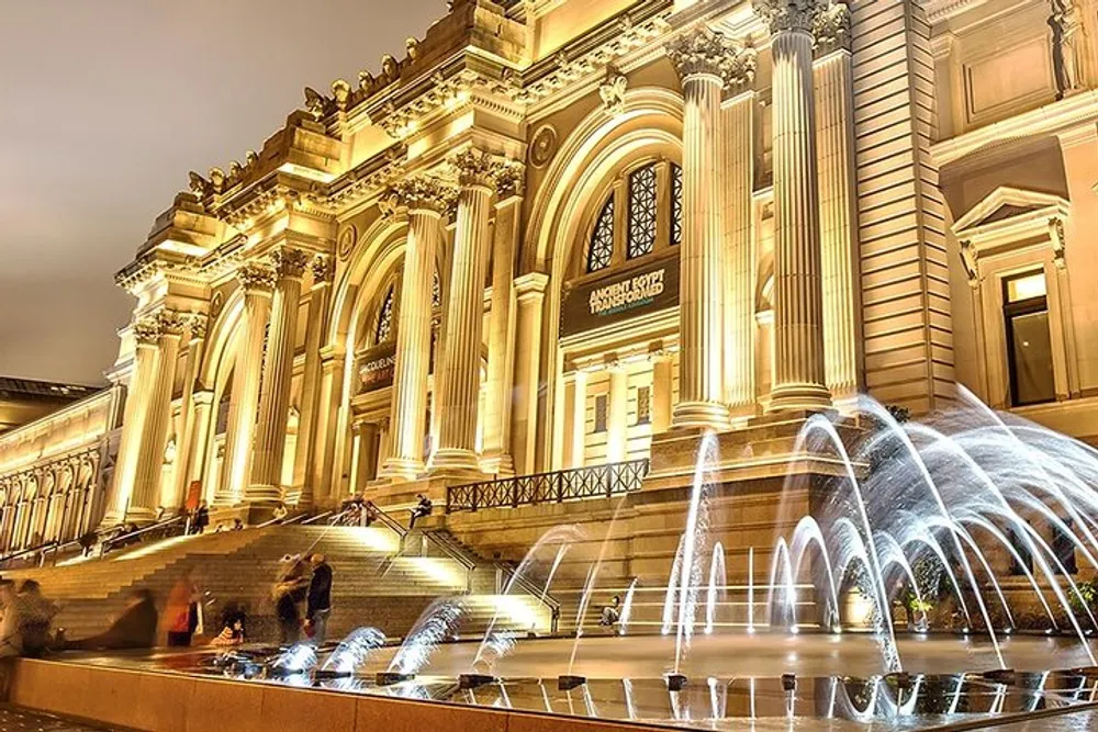 The image shows an illuminated classical building at night with a grand staircase and an elegant fountain in the foreground likely a museum or a similar cultural institution