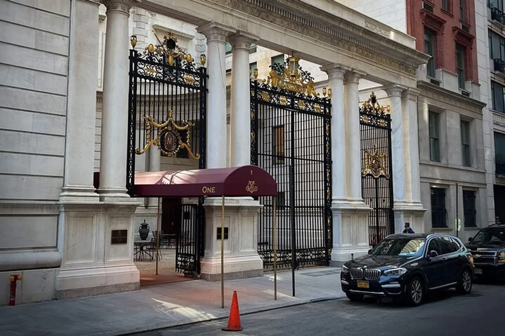 The image shows an elegant entrance gate with ornate gold details on a grand white building marked by a maroon awning that reads ONE and a luxury car parked out front