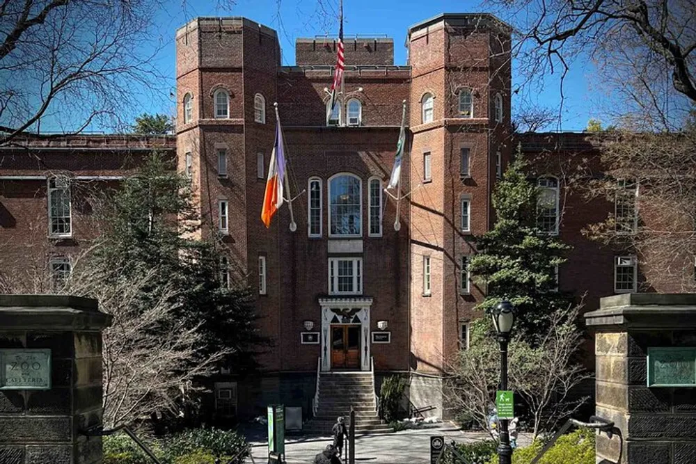 The image shows a brick building with a central entrance flanked by two wings multiple windows and flags hanging above the entrance surrounded by trees under a clear sky