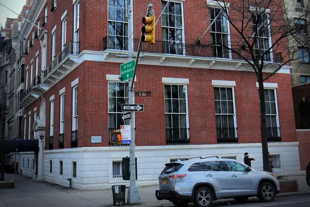 The image shows a red brick building on a street corner with traffic lights street signs a parked car and a pedestrian