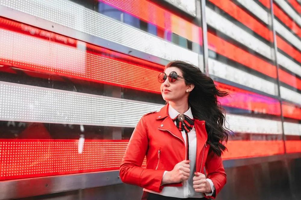 A stylish woman in a red jacket and sunglasses walks confidently in front of a vibrant red LED display