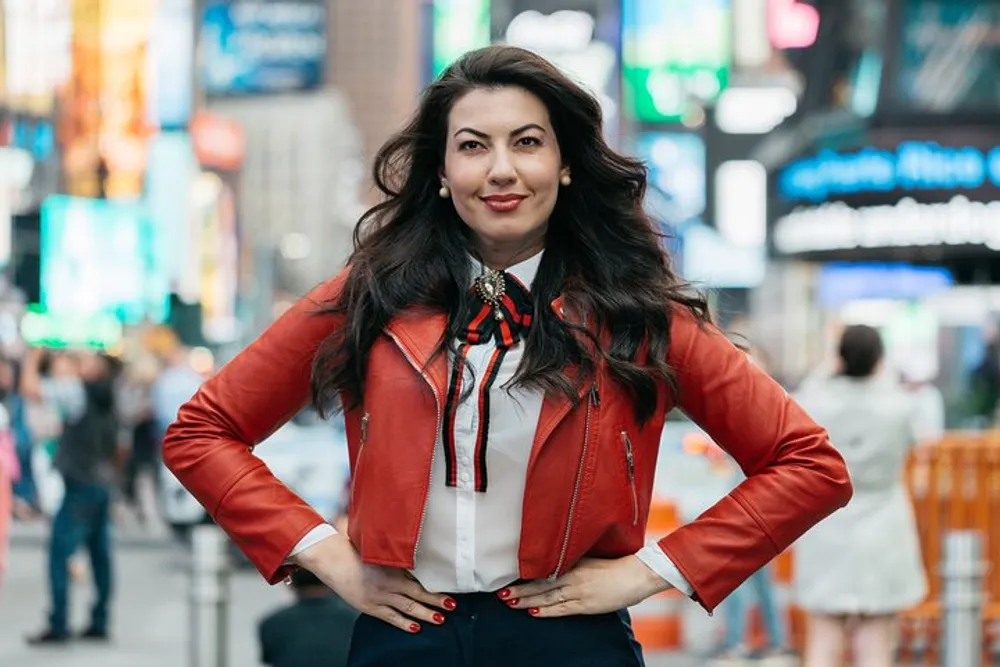 A confident woman in a red leather jacket is standing with her hands on her hips in a bustling urban environment with bright billboards in the background