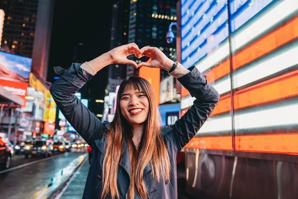 A smiling person is making a heart shape with their hands against the backdrop of a vibrant illuminated city street at night