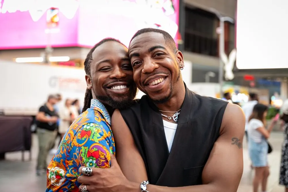 Two happy men are embracing and smiling at the camera in an urban setting that appears to be lively with activity