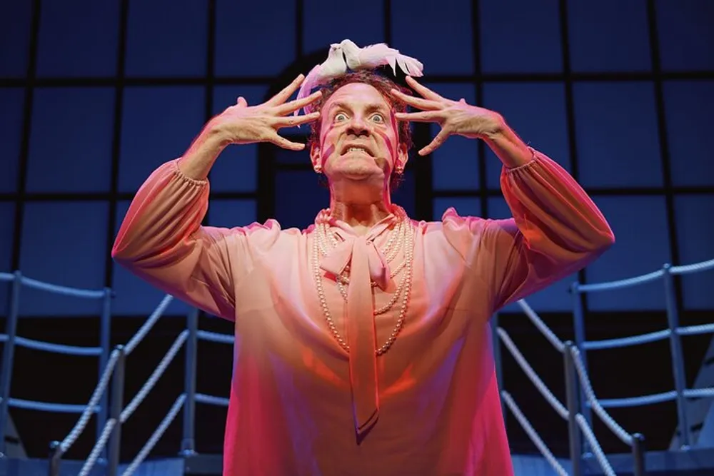 A performer in pink attire is dramatically posing with hands near the face on a stage with a blue grid backdrop