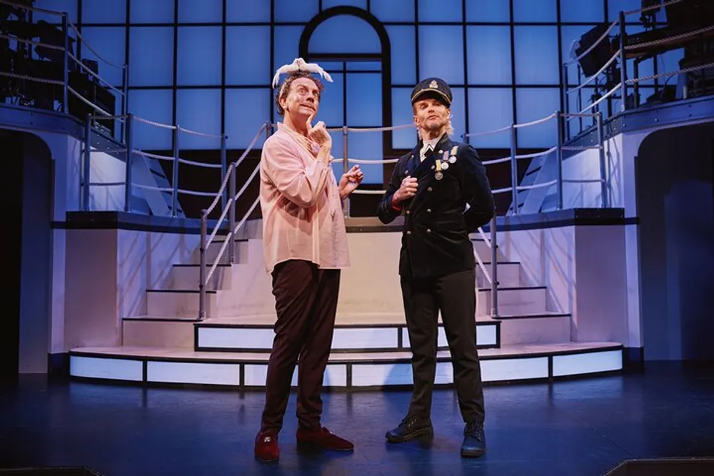 Two actors one dressed in a whimsical costume with a headwrap and the other in a police uniform are performing on stage with a stylized set design in the background