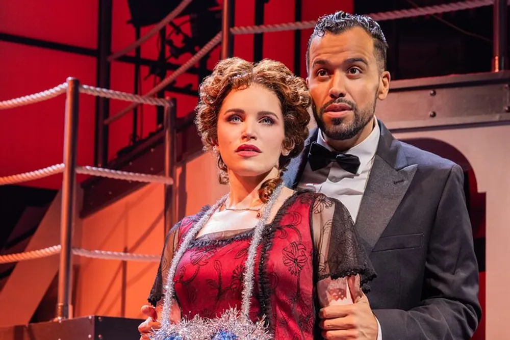 Two actors a woman dressed in a period-style red dress and a man in a tuxedo are performing on a stage set designed to look like an interior with red structures and railing in the background