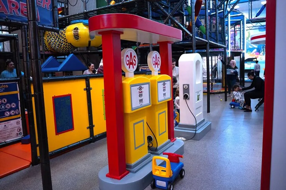 The image shows a colorful play area with a child-sized toy gas station setup alongside a toy car with people in the background enjoying the indoor playground