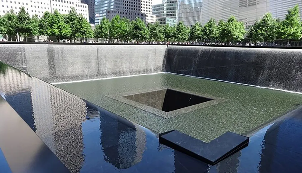 The image depicts one of the twin reflecting pools of the 911 Memorial in New York which are set within the footprints of the original Twin Towers surrounded by trees and skyscrapers