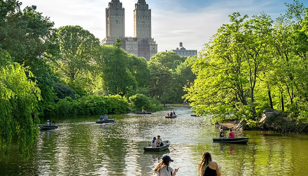 People are enjoying a sunny day with rowboats on a tree-lined lake with a backdrop of towering buildings