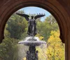 The image shows a statue of an angel with outstretched wings atop a fountain framed by an archway with a backdrop of autumnal trees