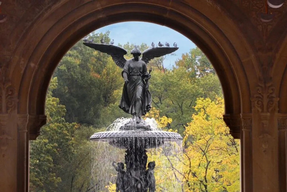 The image shows a statue of an angel with outstretched wings atop a fountain framed by an archway with a backdrop of autumnal trees