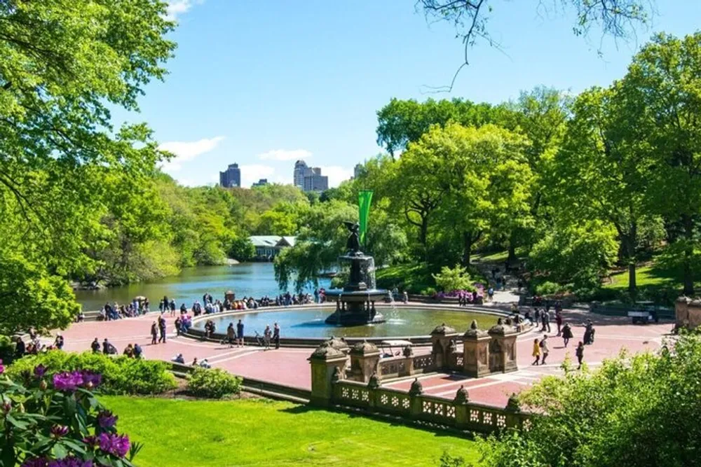 The image shows a vibrant scene at a lush verdant park with people relaxing and walking around a fountain with water and greenery in the background